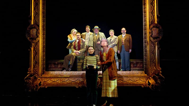 An older, light-skinned woman in house dress and cardigan stands next to a young girl in a striped shirt. Behind them a large, ornate picture frame in which six light-skinned adults stand posed.  
