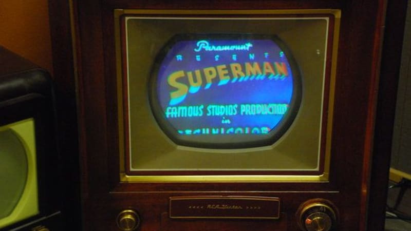 A small, oval screen set inside a dark wooden case. The screen displays a blue background with the word ‘Superman’ over it in reddish-orange letters. 