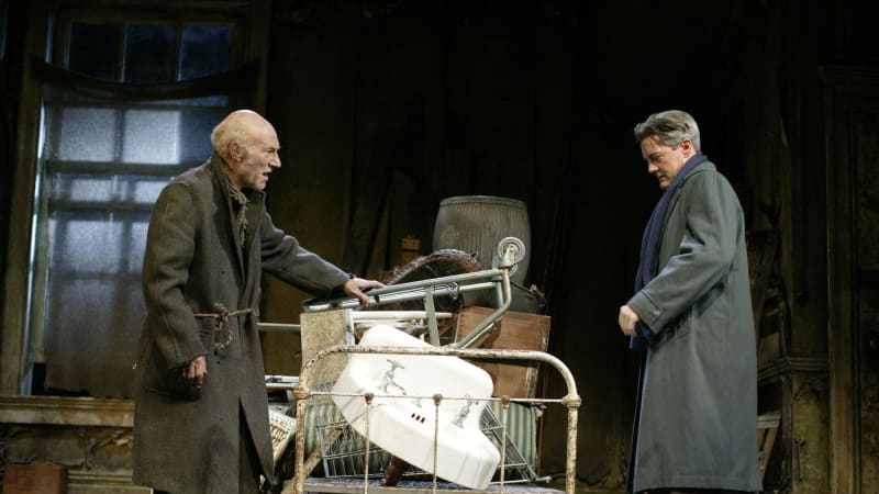 A light-skinned bald man in a tattered coat and shoes extends his hand on top of a pile of objects including a sink and an iron footboard. A light-skinned, well-dressed man stands opposite him. 