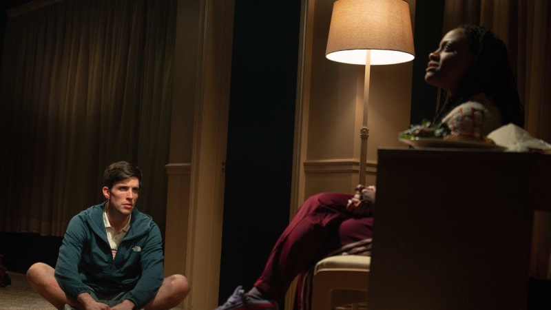 A Black woman in loungewear sits in her desk chair and faces a white man who sits a few feet across from her on the carpeted floor. He wears shorts and has red bug bites on his legs.