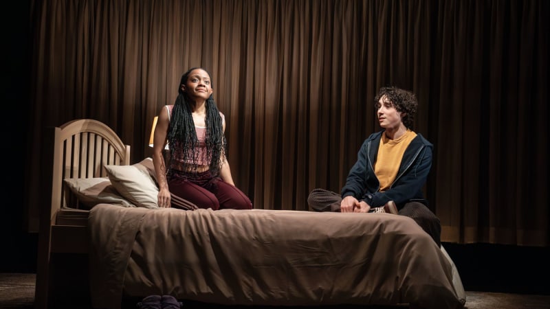 A young Black woman wearing her long braids in pigtails sits on her bed looking up and a young white man with curly hair sits on the bed with her. 