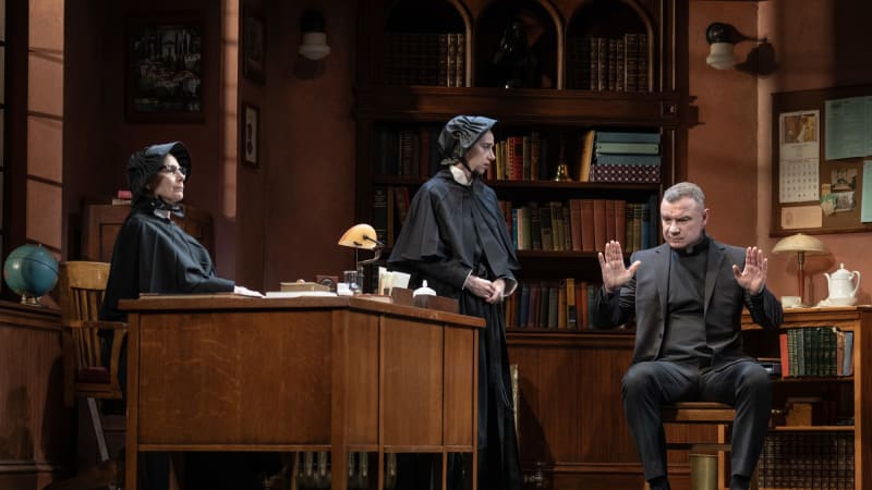 Two nuns and one priest in a principals office. One sister is sitting behind the desk while the other stands. The man looks concerned. 