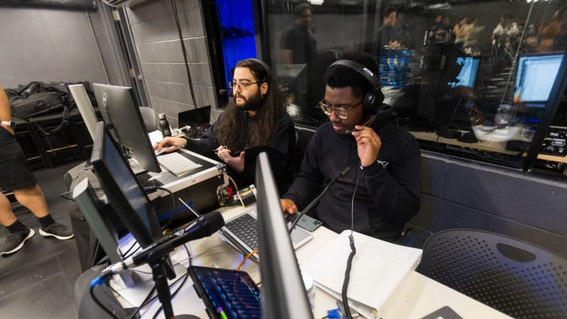 Two TWDP fellows dressed in black wear headsets and sit behind a table full of computers and sound boards.