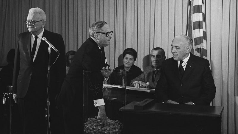 A light-skinned man in a dark suit and dark, rimmed glasses reaches his arm into a tall, clear bucket of capsules as other light-skinned people in formal wear watch. 