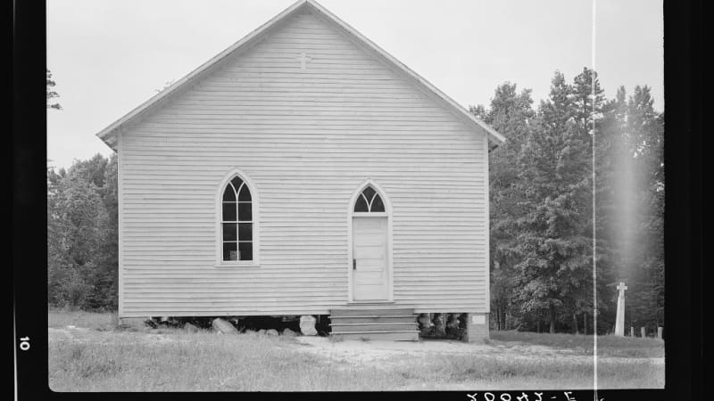 A white building with a peaked roof. On the left side is a window, on the right a set of stairs leading to a door. There is a small cross on the front of the building. 