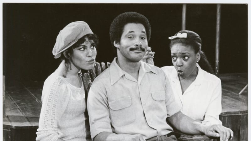 A medium-dark skinned man in workaclothes sits in a rocking chair. Two medium-dark skinned women kneel behind him, making playful facial expressions.