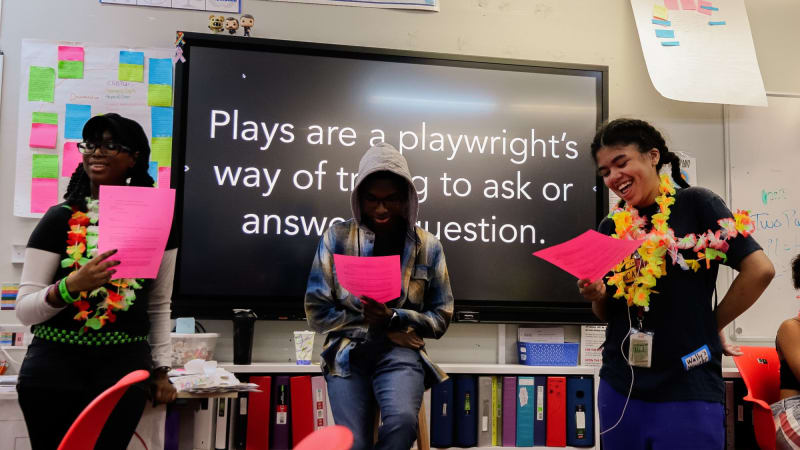 Three smiling students hold pink papers while standing in front of a screen that reads, Plays are a playwright's way of trying to ask or answer a question.