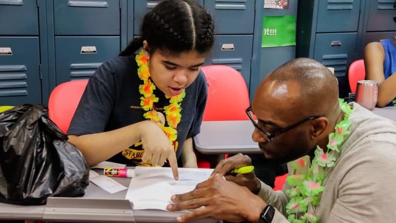 A teacher squats down next to a student's desk as they both look over a piece of paper with highlighters in hand.