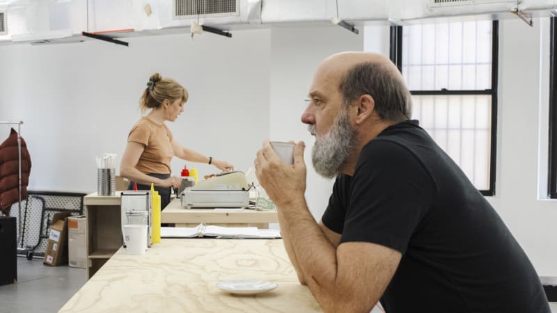 An older man with a full graying beard leans his elbows on a counter as he sips from a coffee cup. Down the counter in the background, a woman with her blonde hair in a claw clip punches numbers into 