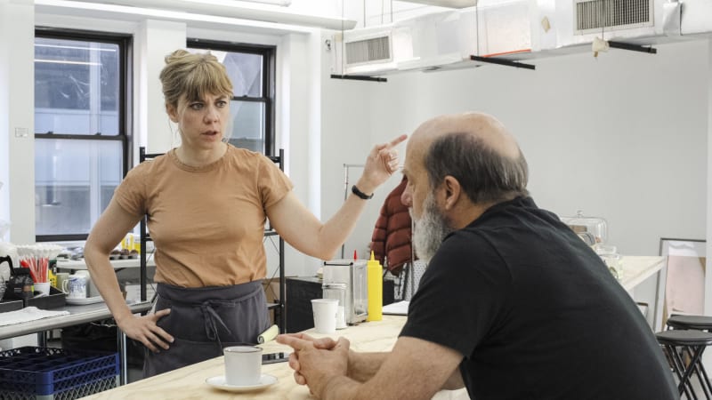 A woman with blonde hair pulled into a clip stands behind a counter with an apron on. She points her finger in a serious gesture while speaking to the older man with a full beard sitting at the counte