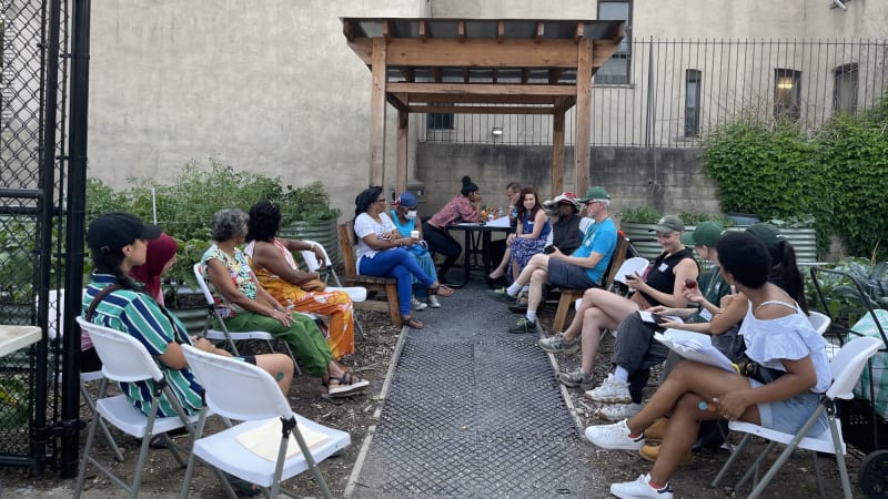 A group of people sit in two rows of chairs facing each other in a community garden. They hold papers and wear name tags.
