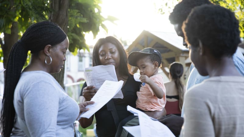 People stand in small groups around a community garden holding scripts. One woman holds both her script and a toddler on her hip.