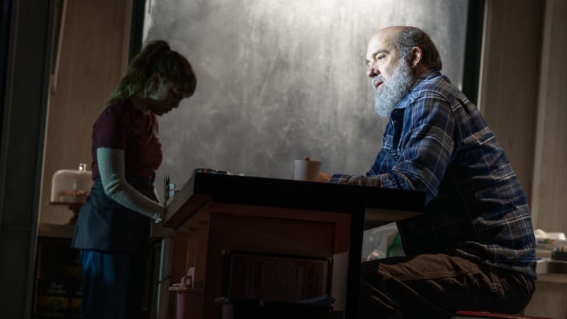 A man with a gray beard wears a blue flannel and sits at the counter of a diner. A soft spotlight illuminates him while silhouetting everything else in the diner.