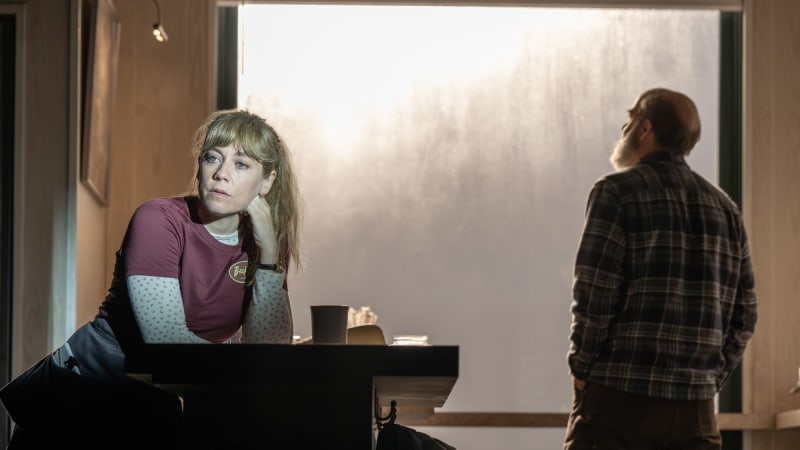 A woman in a blonde ponytail leans on a diner counter with her hand propping up her head. A soft spotlight illuminates her while silhouetting everything else in the diner, including a man looking out 