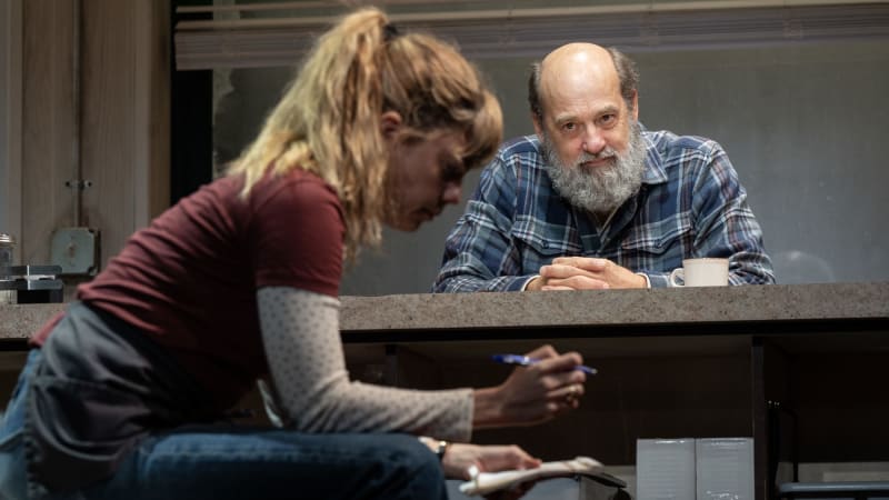 An man with a gray beard leans on a counter as he quietly watches a waitress sitting on a crate writing on a notepad.