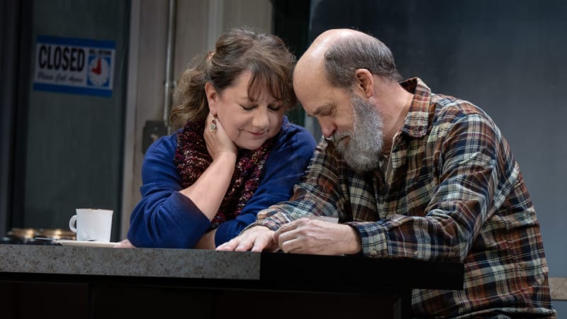 A woman with short brown hair wearing a blue sweater sits next to a man with a gray beard wearing a flannel. They sit shoulder to shoulder with their heads down in a quiet, tender moment.