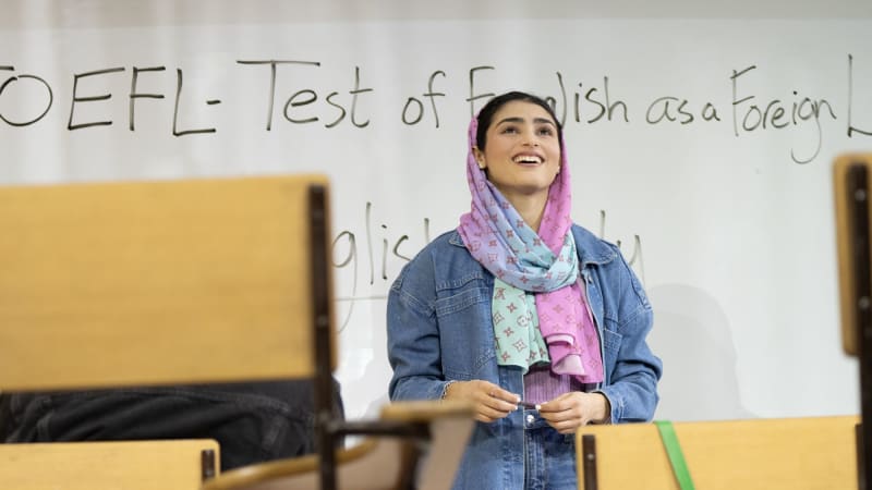 A production image from ENGLISH. A young Iranian woman wearing a pastel pink and blue hijab and a denim jacket smiles and stands in front of a whiteboard in a classroom. 