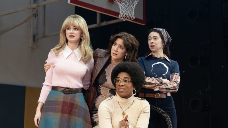 Three concerned women stand close together behind another woman who sits in a folding char in a gymnasium and gestures with her hand like she's making a point.