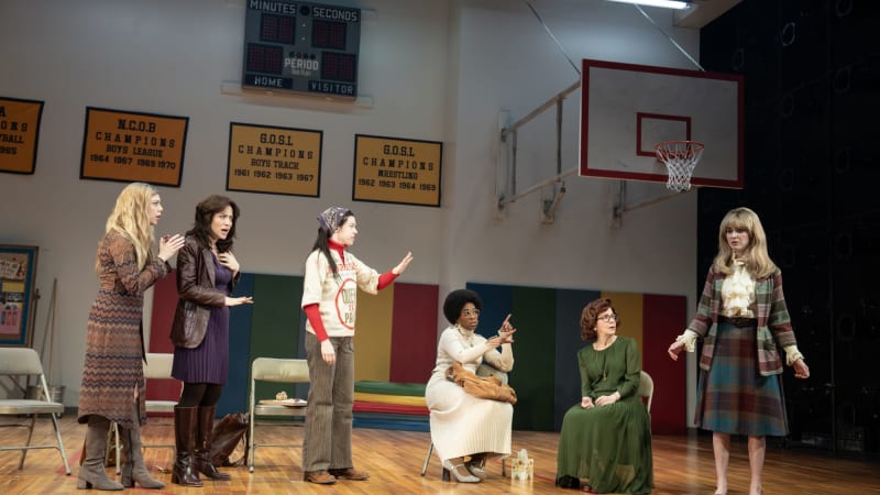 A group of six women in 1970s attire stand and sit around a basket ball. Some  are shocked and some point fingers as they focus their attention on a stunned young woman.
