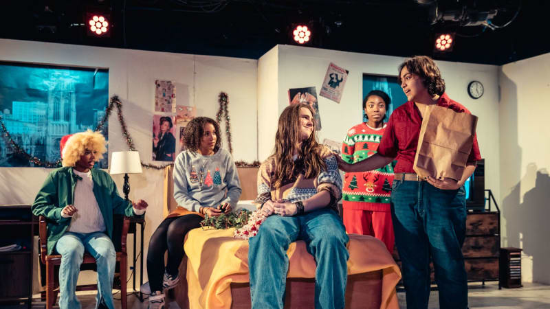 A group of students performing in a play sit and stand around a dorm room set.