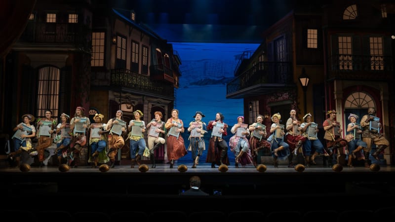 Production image from PIRATES. In a New Orleans street scene, the entire cast lines up at the front of the stage, each person playing a colorful washboard with a bell in unison.
