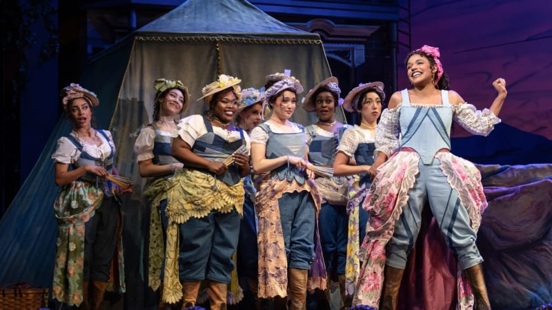 A young woman sings in front of a group of women in a New Orleans beach scene. They all wear matching combinations of denim corsets, colorful bustles and head wraps, and many hold paper fans.