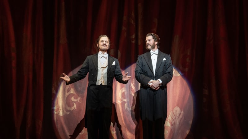 Two men in tailcoat tuxedos and slicked back hair present in front of a red theatre curtain under a round spotlight.
