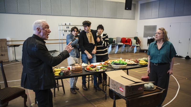 In a rehearsal studio, a man points a finger at a woman in front of a table set for a feast. Behind the table, three young men comically cower together in a clump while watching the tense situation.