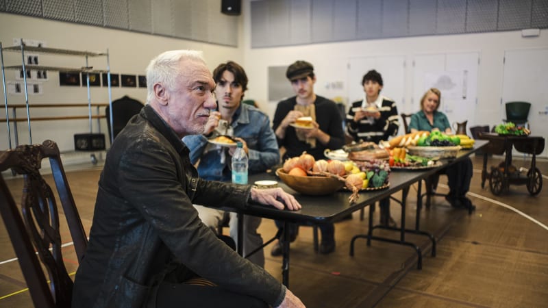 A man in a leather jacket sits at the head of a table set for a feast. Along the table is a row of young men holding plates of food stare at him. A woman watches from the other end of the table.
