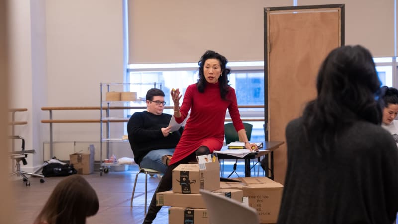 In a rehersal studio an woman holds her hand up speaking besides boxes that are staged like a table at a resturant. Around her are other performers scattered around the space reading their scripts.