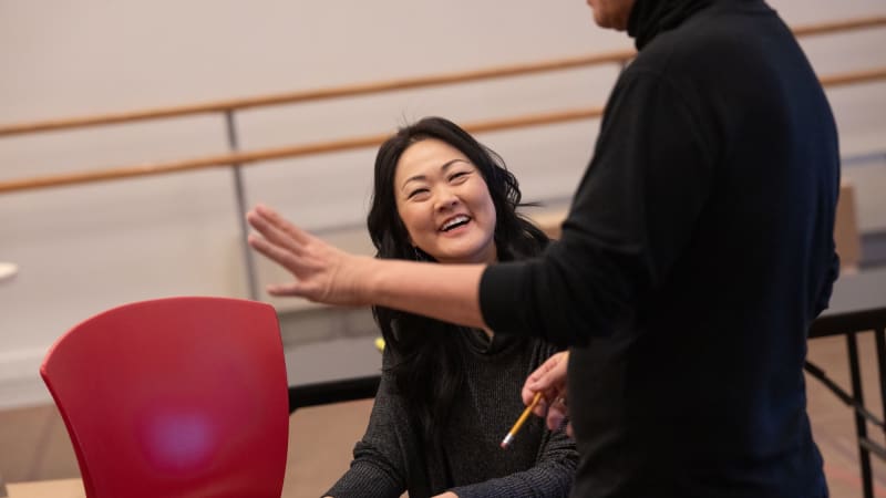 A woman smiling at a man in a rehersal studio space, she is holding a script and pen on a chair as she is listening to him talk.