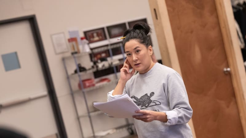 A woman in a rehersal space holding a script and miming as though she's holding a phone and speaking to someone on the phone.