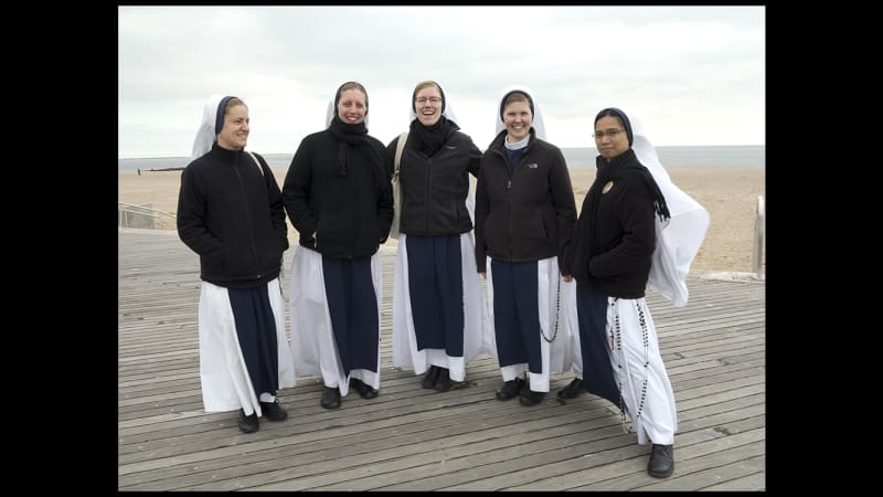 Four light-skinned young women and one medium-skinned young woman in long white and navy habits, white headpieces, and black jackets stand on a boardwalk. Behind them is gray sky, sand, and ocean.
