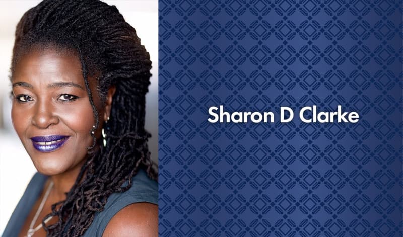 Sharon D Clarke has dark skin, brown eyes, and long black locs. She wears a bright, shiny purple lipstick, silver dangly earrings, black eyeliner, and a blue sleeveless top.