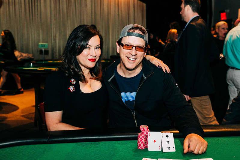 Two people with their arms around each other sit at a poker table and smile. On the green carpeted table are playing cards and red poker chips.