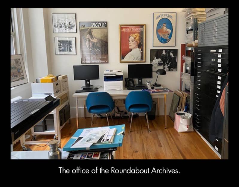 The office of the Roundabout Archives. An office with a long white desk, two blue chairs, and two computer monitors in front of a white wall with six posters on it. 