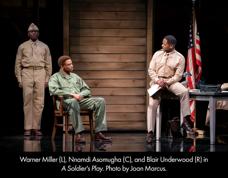 A dark-skinned man in a tan uniform and cap stands at attention at the back left. Center, a dark-skinned man in green fatigues and glasses sits in a chair, speaking to a dark-skinned man in a tan unif