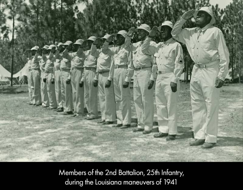 A line of dark-skinned men in light-colored military uniforms and hats. They salute with their right hands.