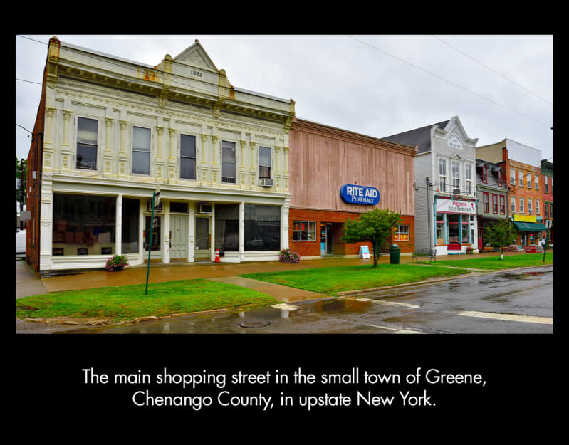 Flat-fronted, two-story buildings including a Rite Aid drugstore and a pizzeria face a wide sidewalk, strip of bright green grass, and a rain-slicked street. 