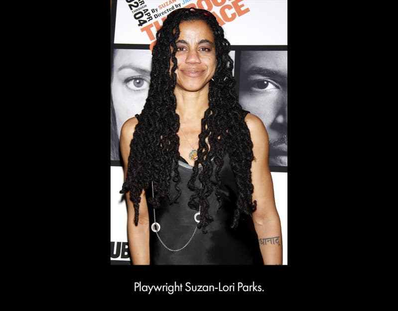 Playwright Suzan-Lori Parks, a medium-brown skinned woman with long, curled locs wears a black dress and stands in front of a poster.