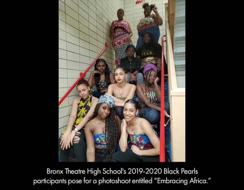 Ten young women with medium or dark skin, wearing shirts made from multi-colored fabric, pose in a school stairwell.