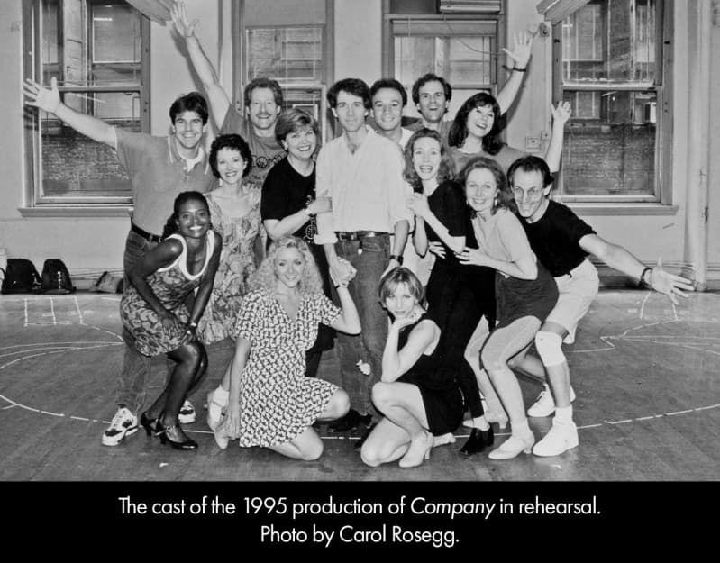 Black and white photo of the 1995 cast of COMPANY in rehearsal. Actors clump together in a group for a photo, some with their arms outstretched.