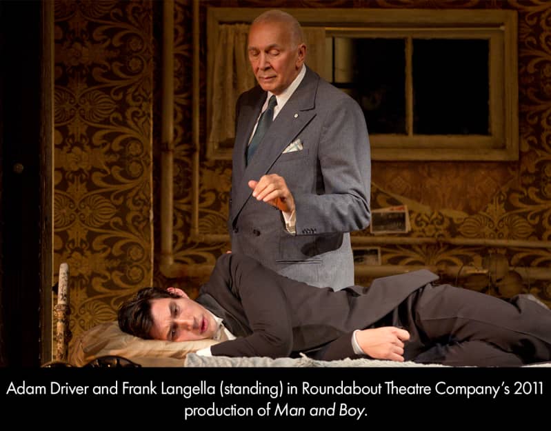 Adam Driver and Frank Langella in MAN AND BOY. A light-skinned young man in a dark gray suit lies on a bed as an older, bald man in a gray suit stands over him, hand lifted to comfort.