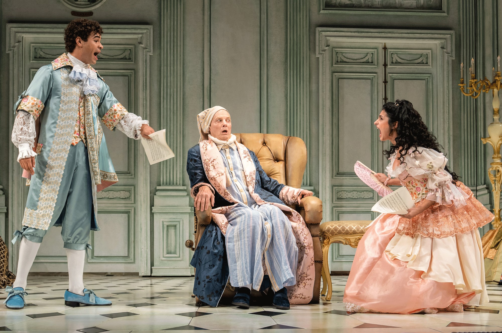 Three actors in colorful period piece costumes on an ornate stage. A woman and man are singing while the third looks on with a surprised expression.