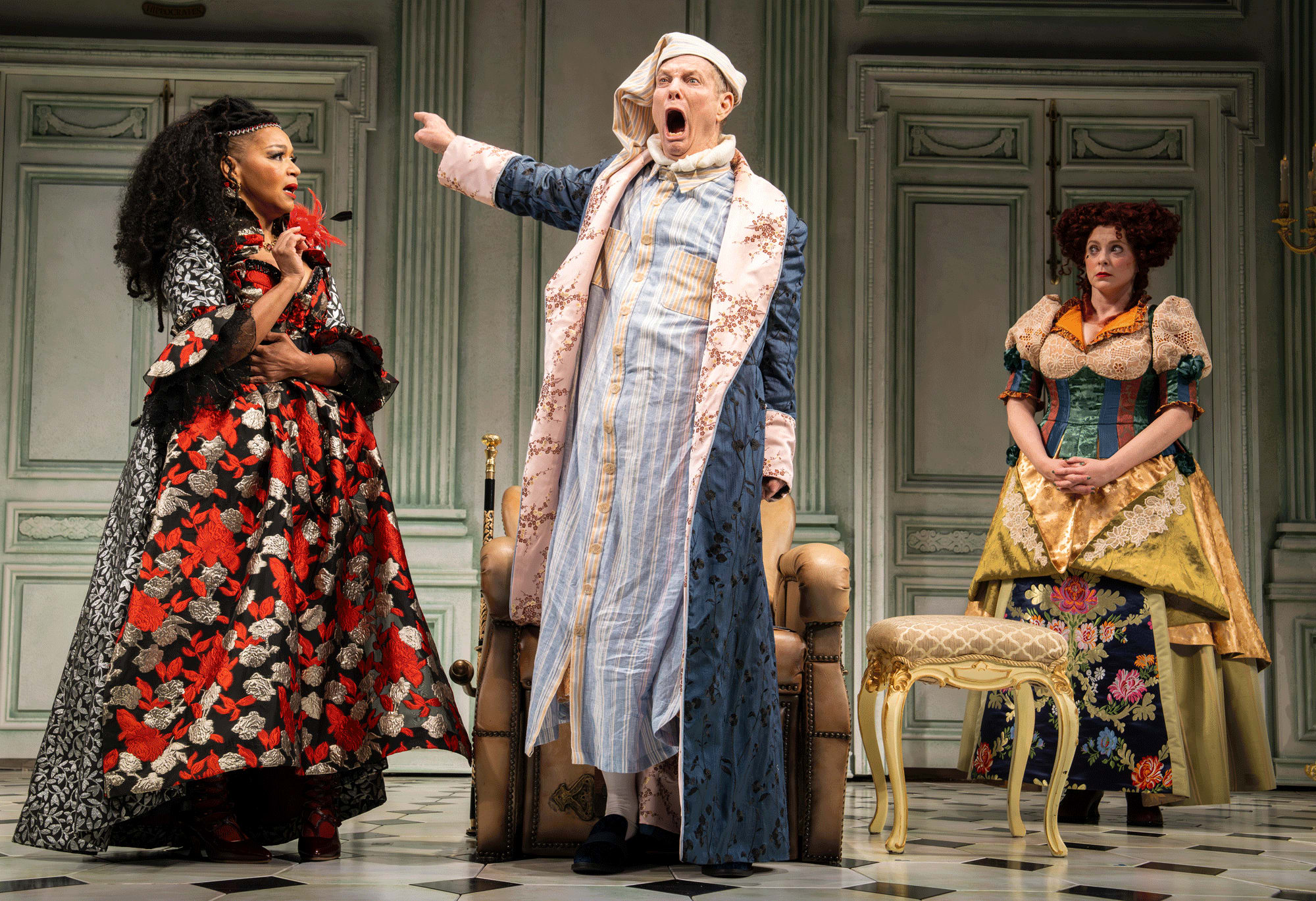 Three actors in colorful period piece costumes on an ornate stage. One appears to be yelling with a finger pointed.