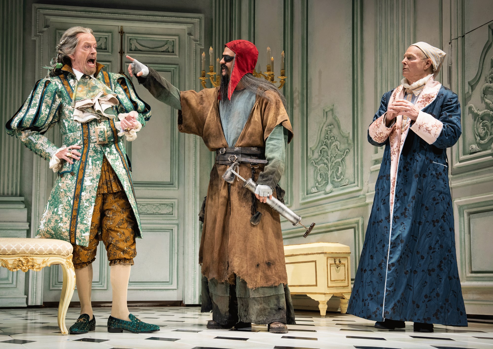Three actors in colorful period piece costumes on an ornate stage. One man points a finger at the other.