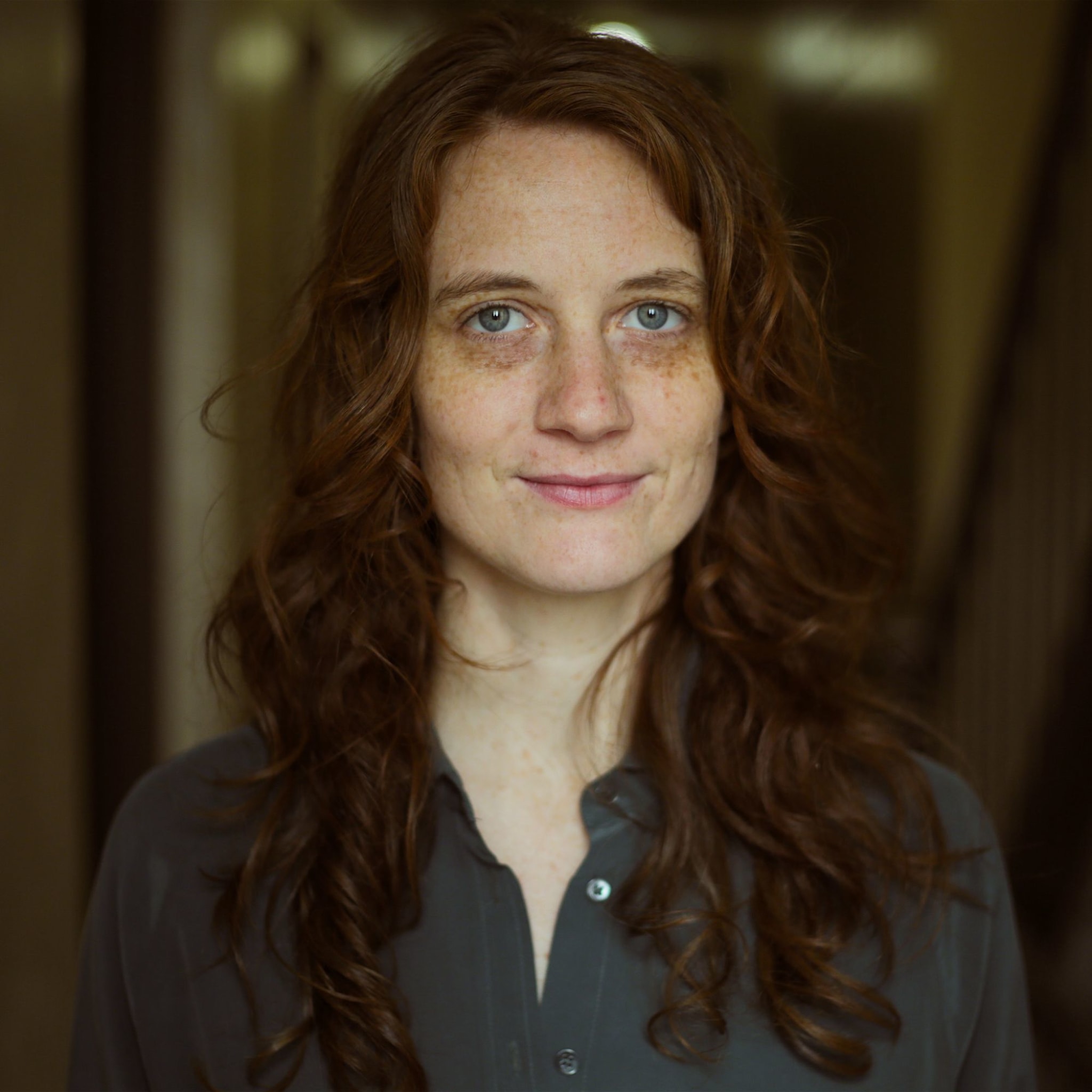 Lee has light skin, freckles, blue-gray eyes, and long, curled brown hair. She wears dark gray henley shirt.