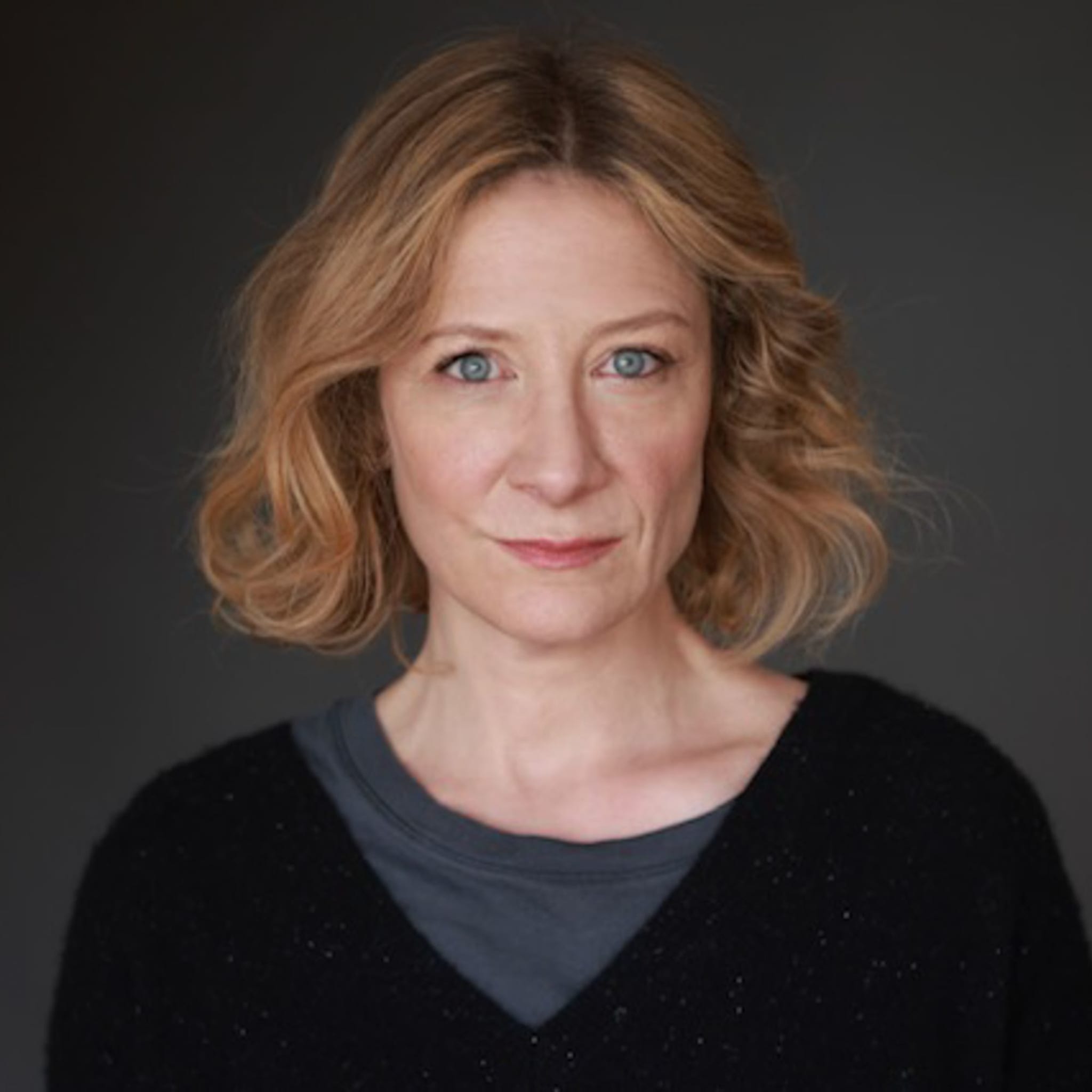 Annie has light skin, large blue eyes, and chin-length, wavy golden blonde hair. She wears a muted dark blue tee under a dark sweater.