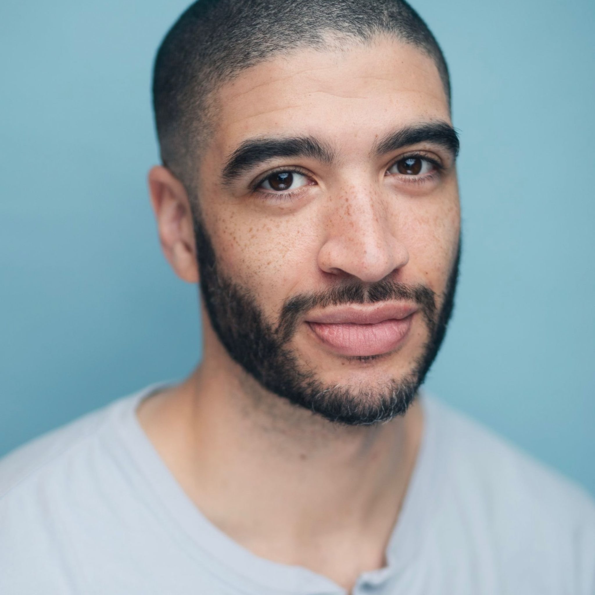 Blake has deep brown eyes, light skin with frecles, dark thick brows, dark facial hair, a closely shaved head, and wears a light gray shirt.