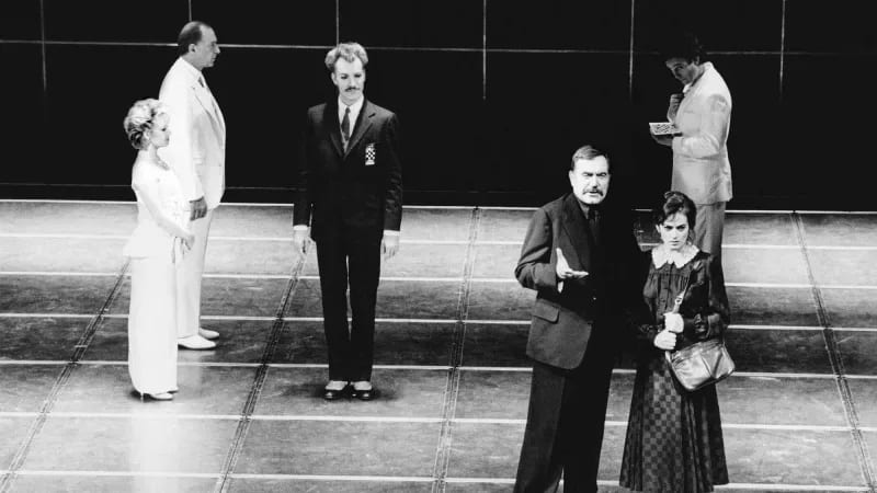 A light skinned, mustached man wearing a suit stands next to a light-skinned woman wearing a long checkered dress. She is clutching a bag on her shoulder and he is gesturing out. To the left, a man and woman wearing all white stand facing a man wearing a suit.